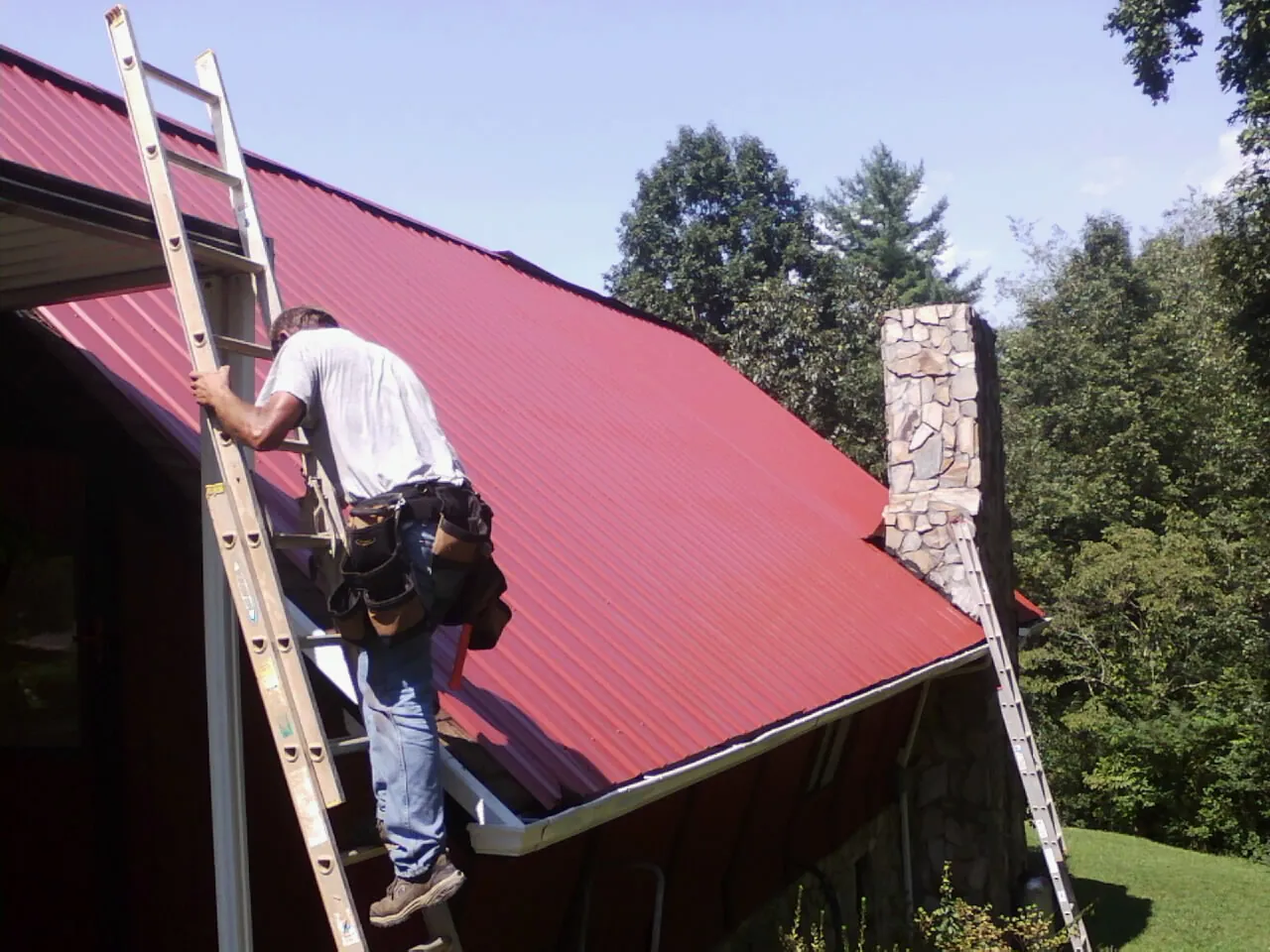 Roofer on a ladder beside a red metal roof and stone chimney — Soffit & Fascia Repair by Roofing Brothers in Fallentimber